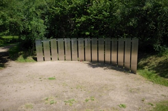 Metallic panels with inscriptions form a memorial set against a backdrop of dense, green foliage. The panels are arranged in a semi-circle on a patch of sandy ground surrounded by grass. Soft shadows are cast on the ground, suggesting a sunny day.