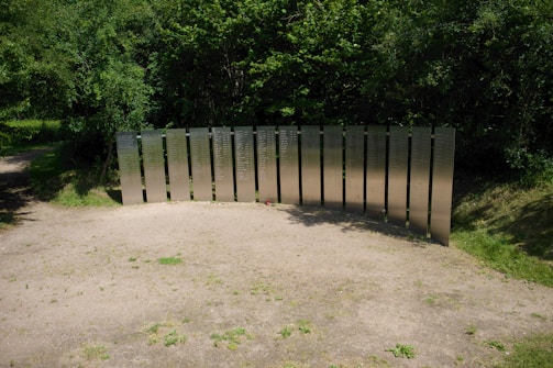 Metallic panels with inscriptions form a memorial set against a backdrop of dense, green foliage. The panels are arranged in a semi-circle on a patch of sandy ground surrounded by grass. Soft shadows are cast on the ground, suggesting a sunny day.