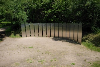 Metallic panels with inscriptions form a memorial set against a backdrop of dense, green foliage. The panels are arranged in a semi-circle on a patch of sandy ground surrounded by grass. Soft shadows are cast on the ground, suggesting a sunny day.