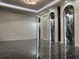 A sleek and modern interior space with polished marble floors and a high ceiling. The room features three tall, arch-shaped panels with decorative lighting and a subtle texture on the walls. A unique ceiling light fixture hangs centrally, adding an elegant touch.