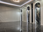 A sleek and modern interior space with polished marble floors and a high ceiling. The room features three tall, arch-shaped panels with decorative lighting and a subtle texture on the walls. A unique ceiling light fixture hangs centrally, adding an elegant touch.