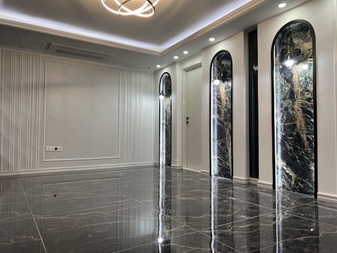 A sleek and modern interior space with polished marble floors and a high ceiling. The room features three tall, arch-shaped panels with decorative lighting and a subtle texture on the walls. A unique ceiling light fixture hangs centrally, adding an elegant touch.