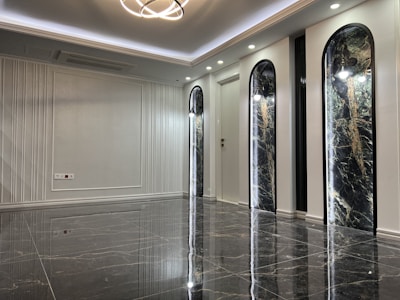 A sleek and modern interior space with polished marble floors and a high ceiling. The room features three tall, arch-shaped panels with decorative lighting and a subtle texture on the walls. A unique ceiling light fixture hangs centrally, adding an elegant touch.