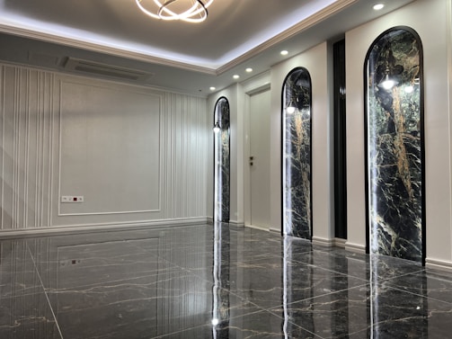 A sleek and modern interior space with polished marble floors and a high ceiling. The room features three tall, arch-shaped panels with decorative lighting and a subtle texture on the walls. A unique ceiling light fixture hangs centrally, adding an elegant touch.