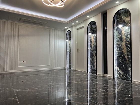 A sleek and modern interior space with polished marble floors and a high ceiling. The room features three tall, arch-shaped panels with decorative lighting and a subtle texture on the walls. A unique ceiling light fixture hangs centrally, adding an elegant touch.