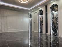 A sleek and modern interior space with polished marble floors and a high ceiling. The room features three tall, arch-shaped panels with decorative lighting and a subtle texture on the walls. A unique ceiling light fixture hangs centrally, adding an elegant touch.