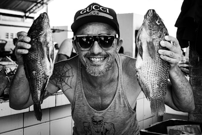 A smiling fishmonger holding a large, glistening fish with the seaside in the background.