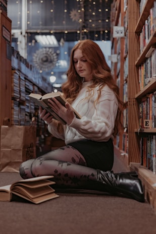 A young woman with long red hair is sitting on the floor of a cozy bookstore or library aisle, engrossed in reading a book. She is wearing a white sweater, a black skirt, patterned tights, and black boots. Surrounding her are tall bookshelves filled with books and some soft, warm lighting creates a serene atmosphere. An open book lays on the floor next to her, and a paper bag can be seen behind her.