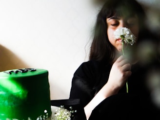 A person with closed eyes holds and smells a white flower, creating a serene and calm atmosphere. A green cake with black decorations sits on a table, partially obscured by flowers at the bottom.