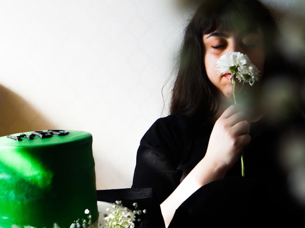 A person with closed eyes holds and smells a white flower, creating a serene and calm atmosphere. A green cake with black decorations sits on a table, partially obscured by flowers at the bottom.