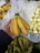 A hand holds a bunch of ripe yellow bananas with a small sticker on one of them. In the background, there are more bananas and some other round, pale-colored fruits. The setting appears to be a market or grocery store with produce displayed in bins.