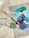 A collection of art supplies including paintbrushes, paint tubes, and a roll of green tape. The surface features a palette of mixed paints in shades of teal, blue, and purple.
