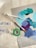 A collection of art supplies including paintbrushes, paint tubes, and a roll of green tape. The surface features a palette of mixed paints in shades of teal, blue, and purple.
