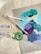 A collection of art supplies including paintbrushes, paint tubes, and a roll of green tape. The surface features a palette of mixed paints in shades of teal, blue, and purple.