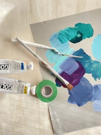 A collection of art supplies including paintbrushes, paint tubes, and a roll of green tape. The surface features a palette of mixed paints in shades of teal, blue, and purple.