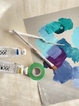 A collection of art supplies including paintbrushes, paint tubes, and a roll of green tape. The surface features a palette of mixed paints in shades of teal, blue, and purple.