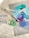 A collection of art supplies is arranged on a surface, including two paint tubes, a roll of green tape, and painting tools such as brushes and palette knives. Several swatches of colorful paint—blue, teal, purple, and turquoise—are visible on a flat surface.
