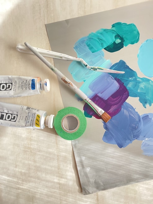 A collection of art supplies is arranged on a surface, including two paint tubes, a roll of green tape, and painting tools such as brushes and palette knives. Several swatches of colorful paint&mdash;blue, teal, purple, and turquoise&mdash;are visible on a flat surface.
