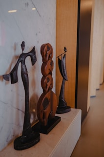 Three abstract sculptures made of dark metal and wood are arranged on a ledge. The figures show artistic human forms with elongated features, showcasing a minimalist and modern design. The background contains a stone wall with visible patterns.