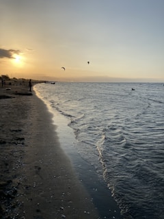 The image captures a serene beach setting during sunset. The sky glows with warm hues of orange and yellow as the sun nears the horizon. Gentle waves ripple towards the sandy shore, where silhouettes of people can be seen enjoying the tranquil atmosphere. In the distance, a few figures are kite surfing, with their kites visible against the sky.