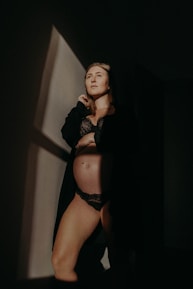 A person is standing in a dimly lit room, wearing black lingerie and a dark robe. The light casts a sharp geometric shadow on the wall, creating a dramatic and intimate atmosphere.