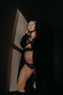A person is standing in a dimly lit room, wearing black lingerie and a dark robe. The light casts a sharp geometric shadow on the wall, creating a dramatic and intimate atmosphere.