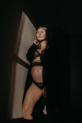 A person is standing in a dimly lit room, wearing black lingerie and a dark robe. The light casts a sharp geometric shadow on the wall, creating a dramatic and intimate atmosphere.