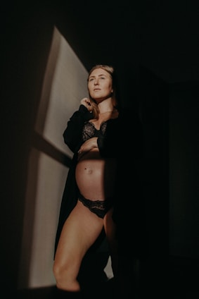A person is standing in a dimly lit room, wearing black lingerie and a dark robe. The light casts a sharp geometric shadow on the wall, creating a dramatic and intimate atmosphere.
