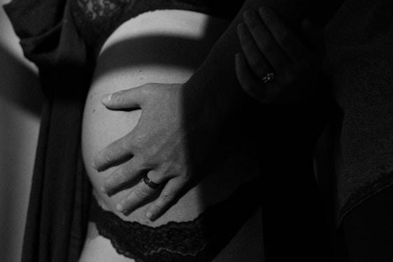 A close-up, black and white image featuring a pregnant belly with two hands gently placed over it, one of which is wearing a ring. The image is intimate and softly lit, highlighting the curves and shadows.