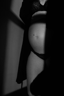A black and white photograph of a pregnant woman wearing lace lingerie. The lighting creates a shadow effect across her body, emphasizing her silhouette. The composition is intimate and artistic, focusing on the curves of her abdomen.