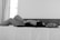 A woman is reclining comfortably on a bed. She appears serene and relaxed, with her eyes closed and hands resting gently on her abdomen. The setting is minimalistic, with a monochromatic black and white tone enhancing the peaceful ambiance.