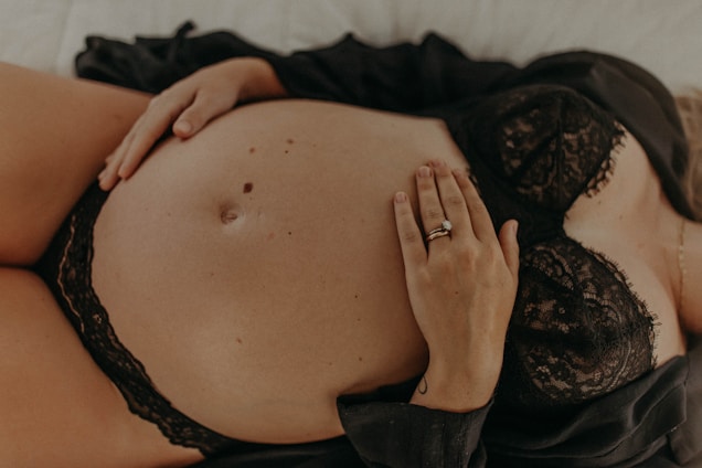 A close-up image of a pregnant woman lying down. She is wearing a black lace bra, and her hand with a wedding ring rests on her bare belly. Another hand is placed below the belly, also seeming to gently touch it. The overall image has a warm, intimate feel.