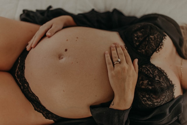 A close-up image of a pregnant woman lying down. She is wearing a black lace bra, and her hand with a wedding ring rests on her bare belly. Another hand is placed below the belly, also seeming to gently touch it. The overall image has a warm, intimate feel.