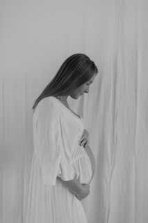 A black and white portrait of a pregnant woman in a profile view, gently holding her belly. She is wearing a long, flowing dress and standing against a plain, softly textured background.