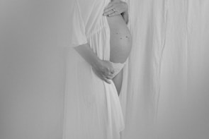 A black and white photograph of a pregnant person gently holding their belly. The individual is partially covered in a sheer, flowing fabric, creating a serene and tender atmosphere. The focus is on the belly, conveying a sense of anticipation and motherhood.