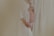 A close-up view of a pregnant woman gently holding her belly, partially covered by a light, sheer robe. Her left hand is placed above her belly, while her right hand supports from below, suggesting a tender, protective gesture. The background consists of a soft, cream-colored fabric, contributing to the serene and intimate atmosphere.