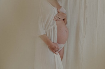 A close-up view of a pregnant woman gently holding her belly, partially covered by a light, sheer robe. Her left hand is placed above her belly, while her right hand supports from below, suggesting a tender, protective gesture. The background consists of a soft, cream-colored fabric, contributing to the serene and intimate atmosphere.