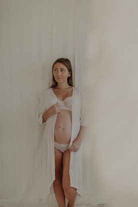 A woman stands against a light-colored background, wearing a lace undergarment and an open robe. Her expression is serene, with her gaze directed slightly to the side.
