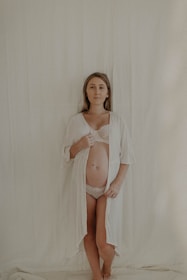 A woman stands against a plain, light-colored backdrop, wearing a sheer white robe open to reveal her pregnant belly. Her expression is calm and serene.