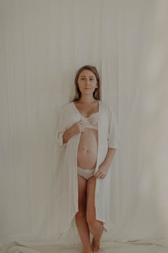 A woman stands against a plain, light-colored backdrop, wearing a sheer white robe open to reveal her pregnant belly. Her expression is calm and serene.