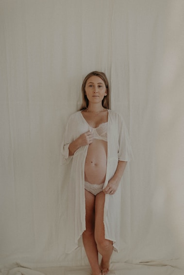 A woman stands against a plain, light-colored backdrop, wearing a sheer white robe open to reveal her pregnant belly. Her expression is calm and serene.