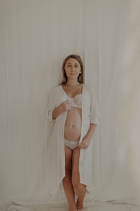 A woman stands against a plain, light-colored backdrop, wearing a sheer white robe open to reveal her pregnant belly. Her expression is calm and serene.