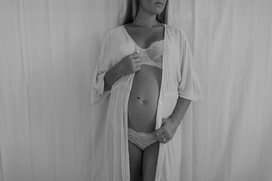 Monochrome photograph of a pregnant woman wearing lace lingerie and an open front robe. The woman is holding the robe with both hands and is standing in front of a plain backdrop.