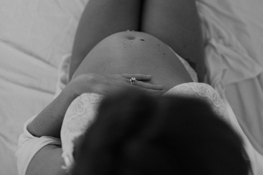 A black and white photograph capturing an intimate angle of a pregnant woman's belly. Her hands rest gently on her stomach, with a visible wedding ring. The fabric she is wearing appears soft and delicate, enhancing the serene atmosphere.