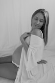 A pregnant woman poses gracefully, draped in a soft white robe, sitting on a similarly colored fabric. Her eyes are gently closed, conveying a serene and peaceful expression.