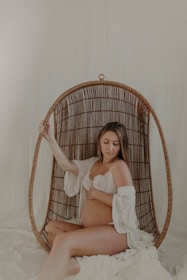 A woman sits in a wicker hanging chair, gently cradling her pregnant belly, wearing a lace bra and a loosely draped white garment. The setting is softly lit, creating a serene and intimate atmosphere.