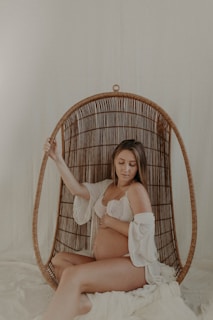 A woman sits in a wicker hanging chair, gently cradling her pregnant belly, wearing a lace bra and a loosely draped white garment. The setting is softly lit, creating a serene and intimate atmosphere.