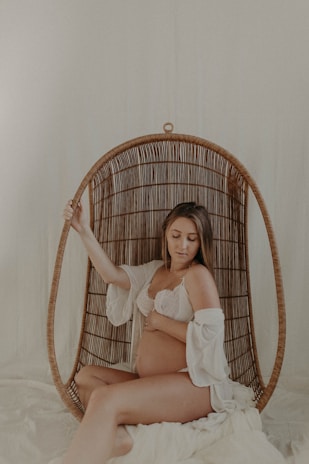 A woman sits in a wicker hanging chair, gently cradling her pregnant belly, wearing a lace bra and a loosely draped white garment. The setting is softly lit, creating a serene and intimate atmosphere.