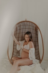 A pregnant woman sits comfortably in a woven hanging chair with a soft, serene expression. She is dressed in a white lace bra and an open robe, gently cradling her belly. The setting exudes a calm, intimate atmosphere with neutral and warm tones.