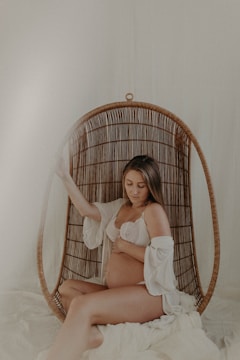 A pregnant woman sits comfortably in a woven hanging chair with a soft, serene expression. She is dressed in a white lace bra and an open robe, gently cradling her belly. The setting exudes a calm, intimate atmosphere with neutral and warm tones.